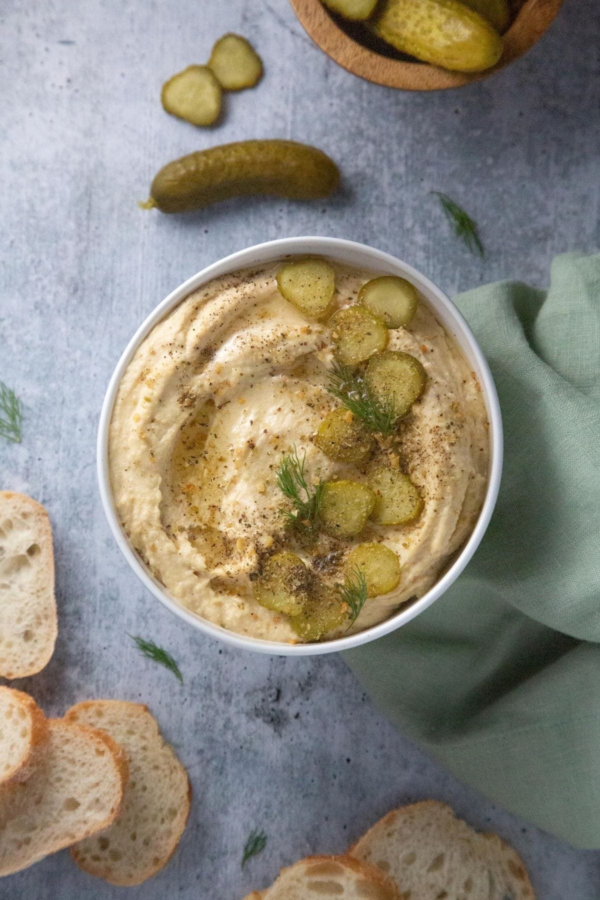 A bowl of hummus with pickle slices and dill on top place on a grey table surrounded by crostinis.