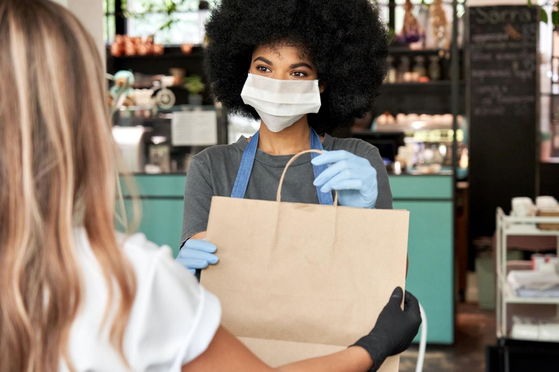 An woman wearing a mask hands a paper takeout bag to customer.