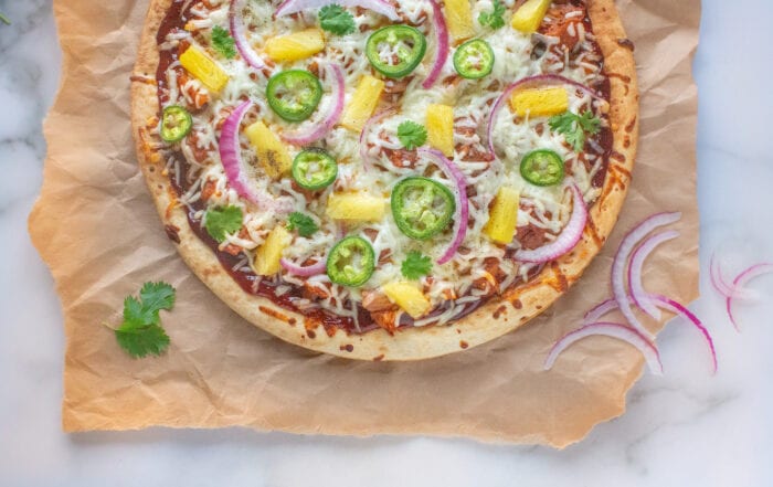 Picture of a pizza topped with barbecue sauce, plant-based meat substitute jackfruit, vegan mozzarella, sliced jalapeños, pineapples chunks, and sliced red onion