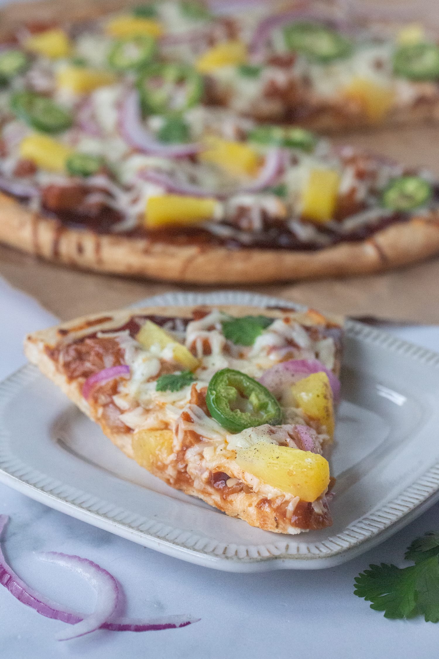 Picture of a pizza topped with barbecue sauce, plant-based meat substitute jackfruit, vegan mozzarella, sliced jalapeños, pineapples chunks, and sliced red onion