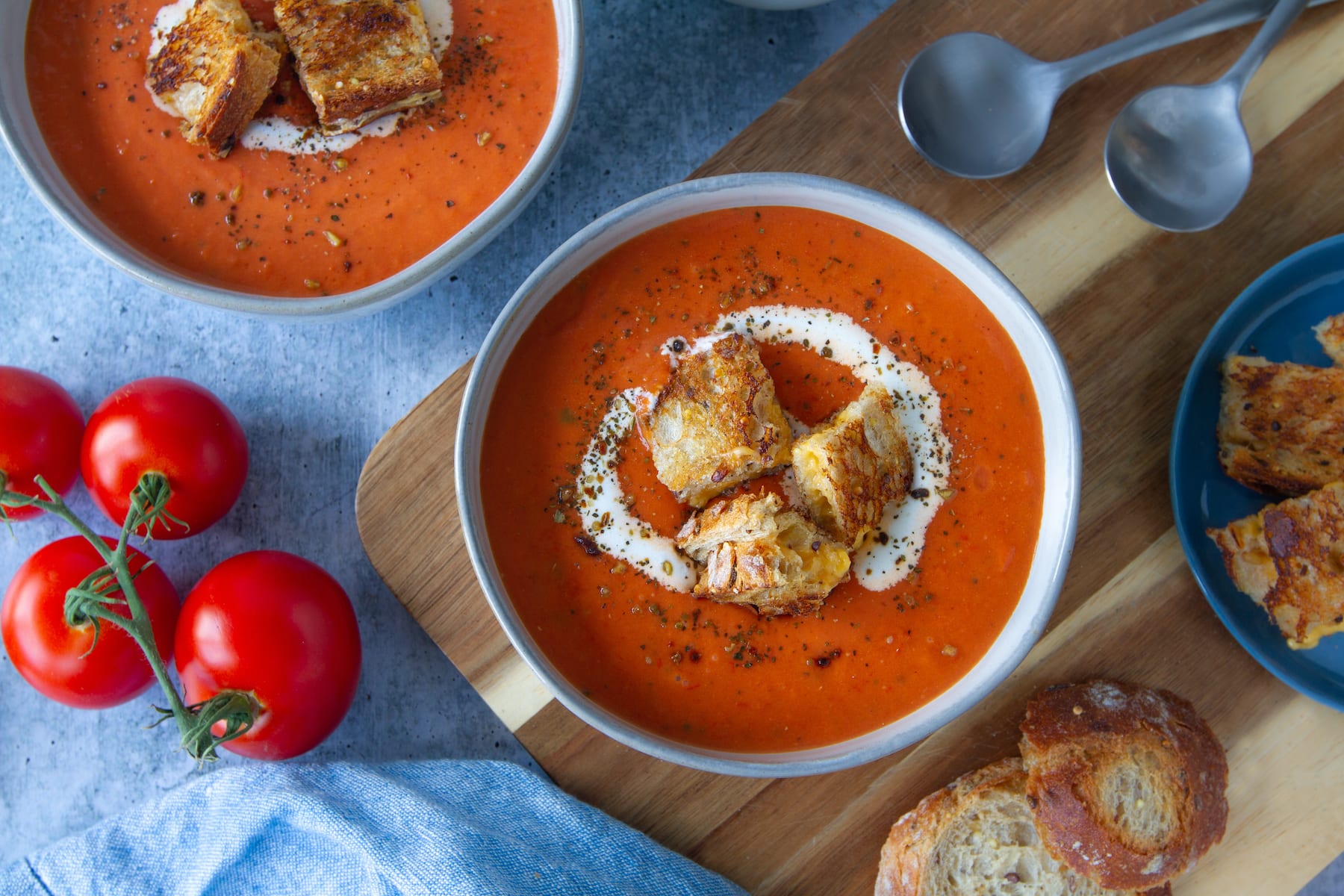 ImaAn overhead shot of two big bowls of tomato soup, drizzled with vegan cream and topped with rustic grilled cheese croutons. Soup spoons and extra tomatoes rest next to soup.ge