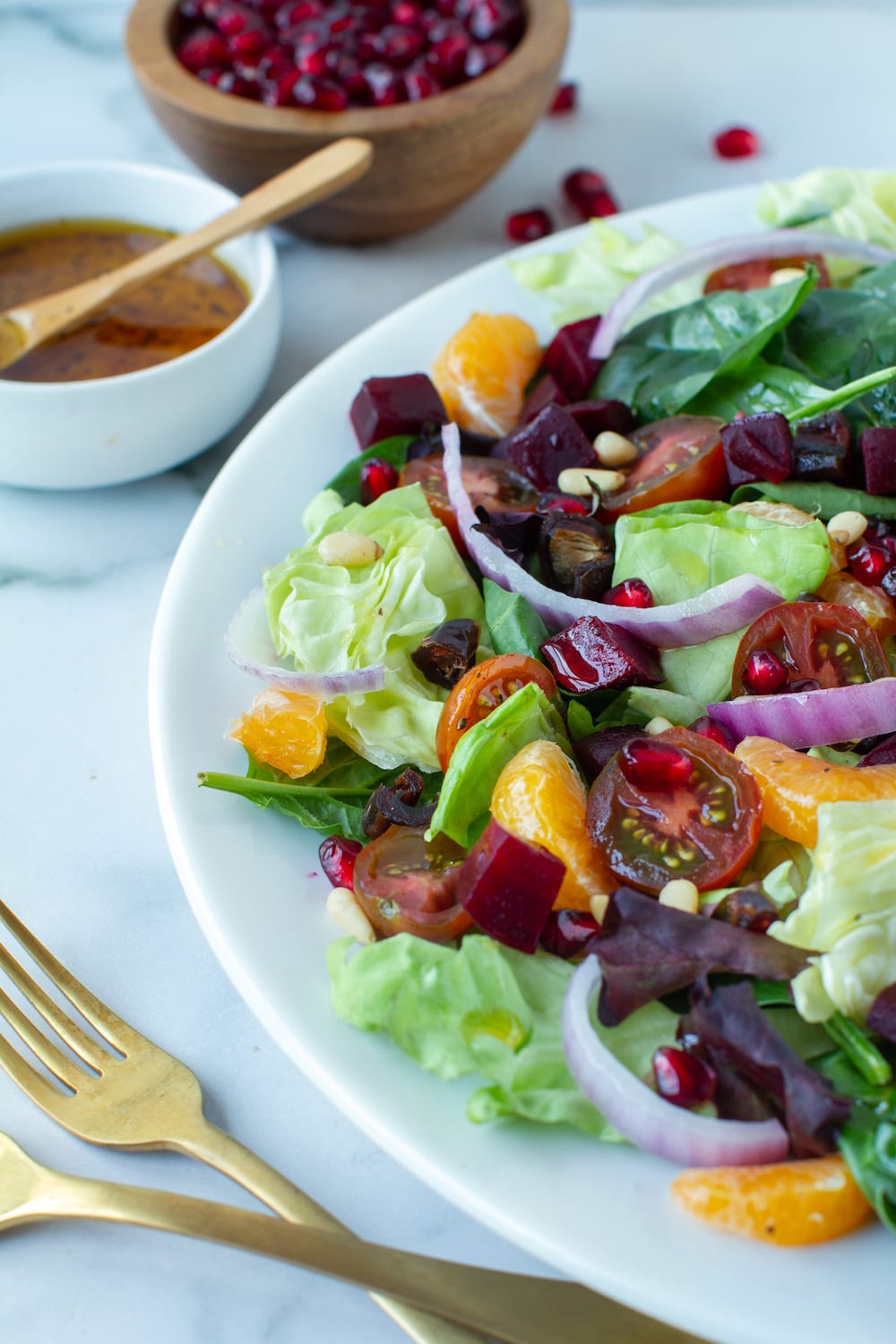 A bed of spinach and lettuce topped with pomegranates, pine nuts, clementine segments, julienned red onions and chopped beets. Salad dressing sits off to the side.