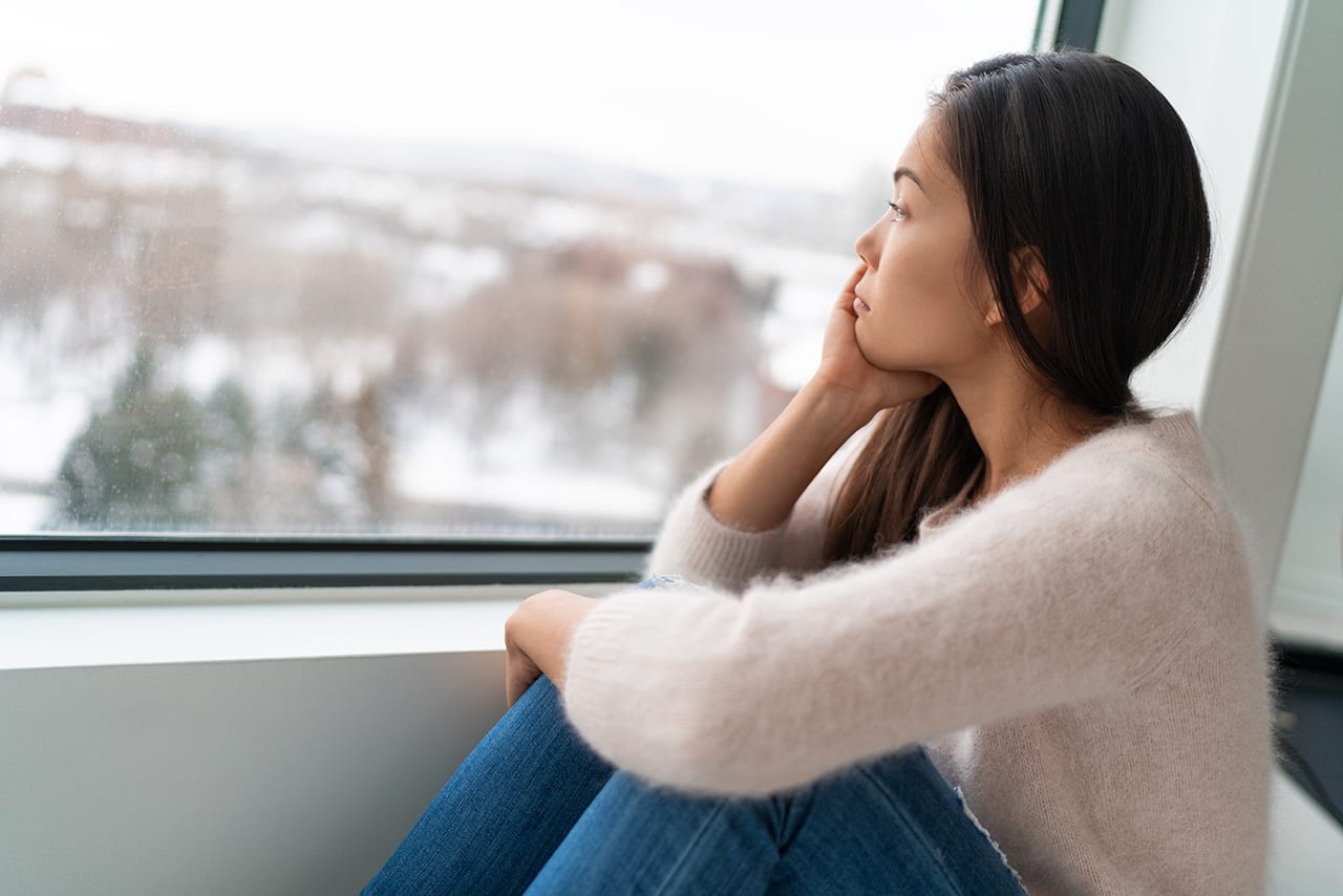 A young woman in a white sweater and blue jeans sits on a large windowsill. She cups her hand around her face and looks out onto a blurry, snowy scene. Her expression is troubled.