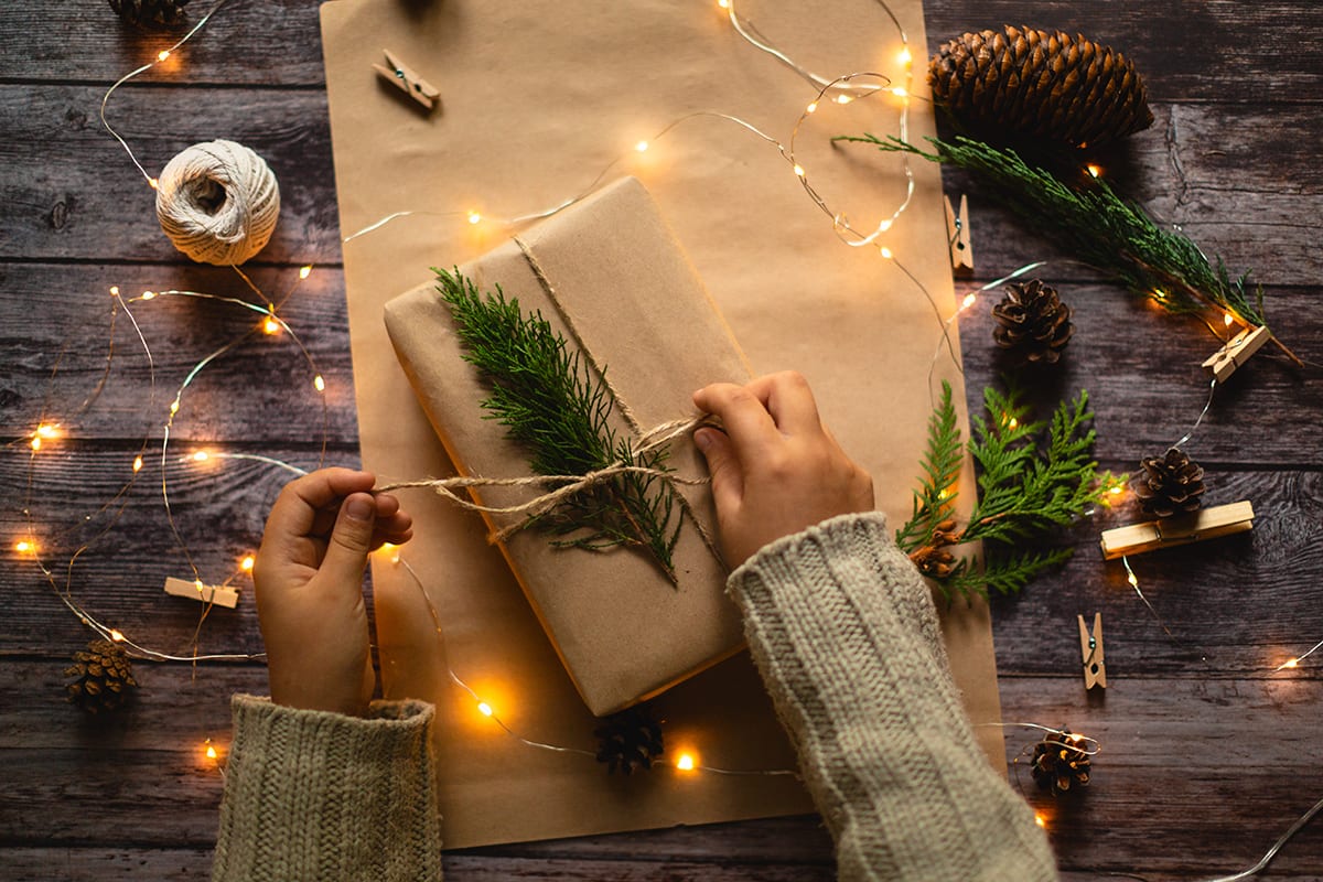 Two hands are tying a piece of light brown twine around a present wrapped in recyclable brown paper. The present is also decorated with part of a pine branch. The hands and the present are surrounded by pine cones, more pieces of pine, and soft white twinkle lights.