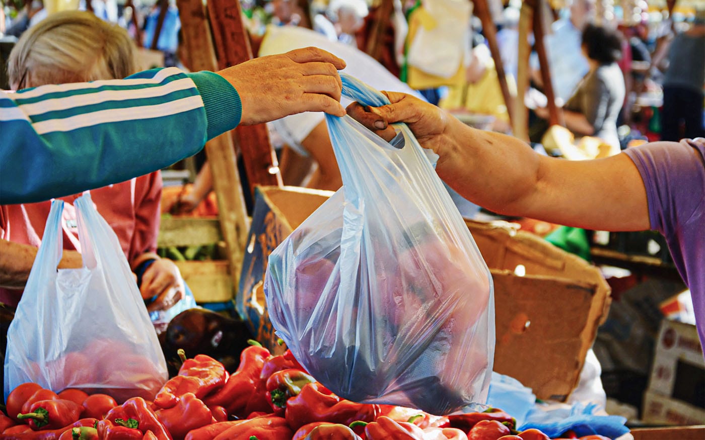 In a busy outdoor market, one person takes a plastic bag full of fruits and vegetables from the seller.