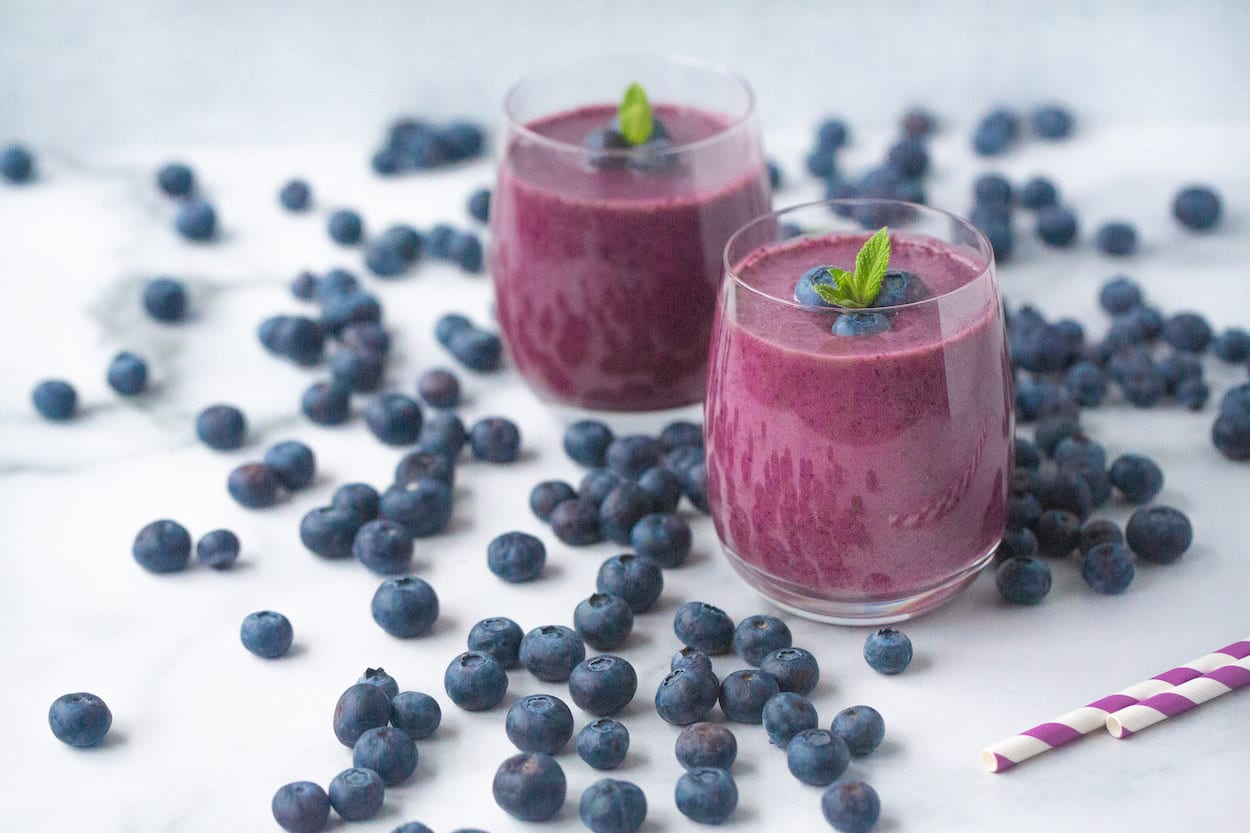 Two ready-to-enjoy Banana Blueberry Smoothies sit in the middle of hundreds of fresh blueberries.