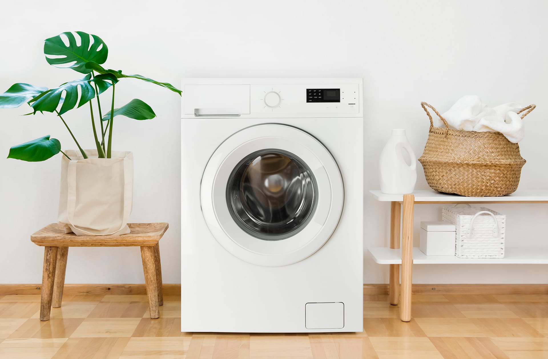 A large white washing machine sits on a wooden floor in between a large palm plant on the left and a small wooden shelf on the right. A wicker basket with white towels sits on the shelf, and next to it is a plain, all-white bottle of laundry detergent.