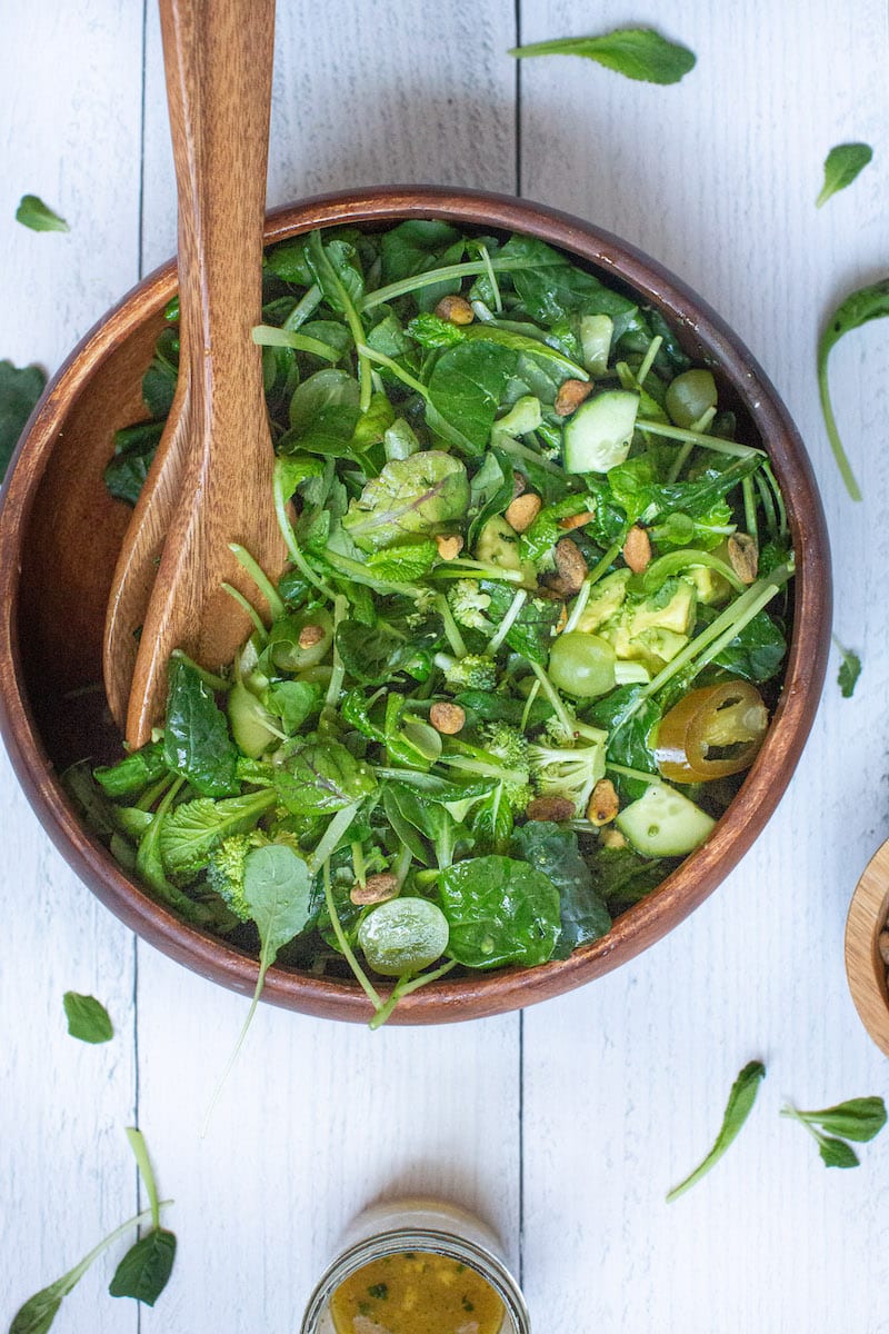 A bowl of fresh green salad ingredients assembled with a jar of dressing and small, wispy bits of spinach and mixed greens strewn about.